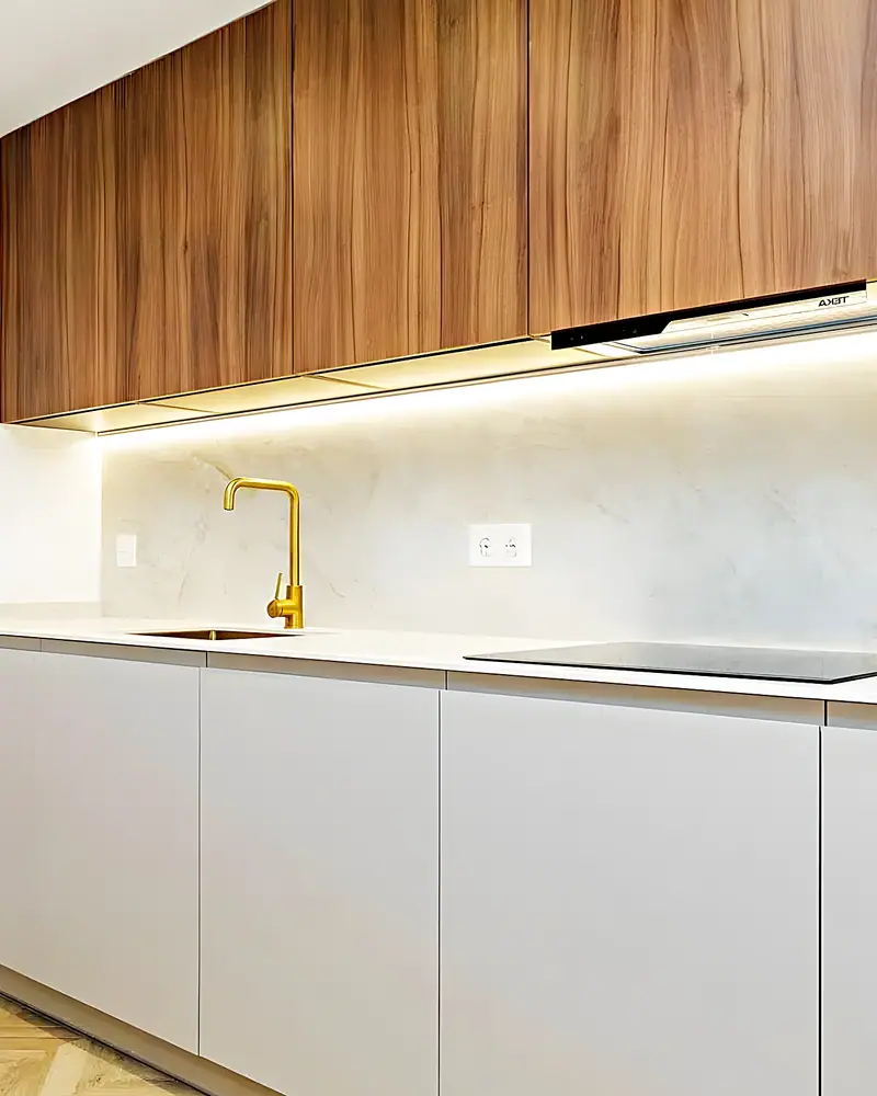 Encimera clara con grifo dorado, frente de piedra y muebles altos de nogal con luz LED